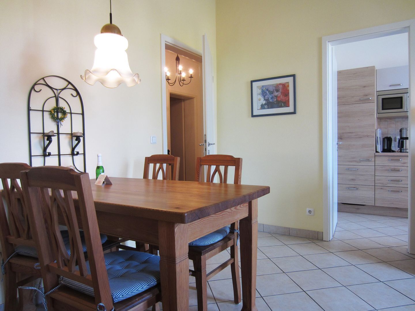 Un coin repas avec une table et des chaises en bois, un lustre et une cuisine visible à travers une porte ouverte.