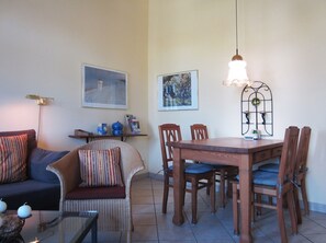 Un salon comprenant un canapé, un fauteuil en osier, une table à manger avec des chaises et deux tableaux accrochés au mur.