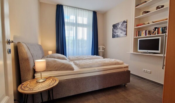 A bedroom with a bed, a bedside lamp, a round table with a compass design, a TV on a stand, and a bookshelf.