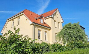 Ein zweistöckiges Gebäude mit roten Ziegeln auf dem Dach, zahlreichen Fenstern und üppiger Bepflanzung im Vordergrund.