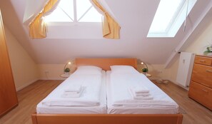Un letto matrimoniale con biancheria da letto e asciugamani bianchi, una testiera in legno, comodini con lampade, un termosifone e una cassettiera di legno.