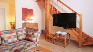 Una sala de estar con una escalera de madera, un sofá, un televisor sobre un mueble bajo y una lámpara.