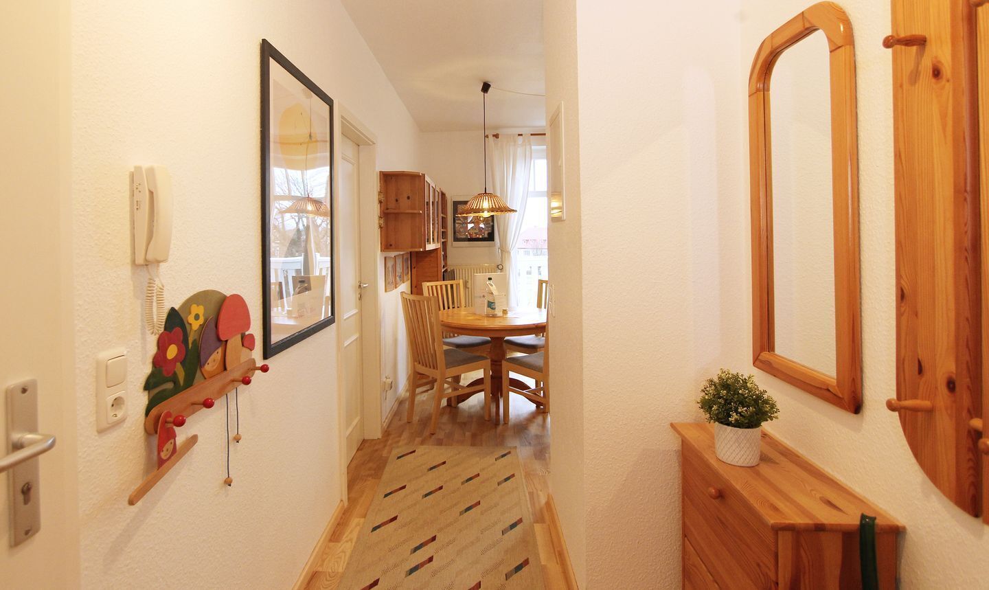 Ein schmaler Flur mit Holzboden, ein Essbereich mit Holztisch und -stühlen sowie ein kleiner Blumentopf auf einer Holzkommode.