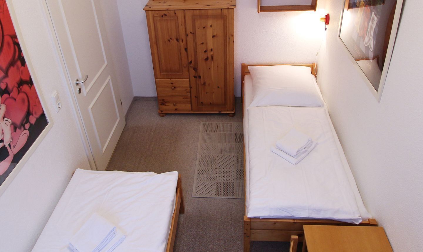 Ein kleines, schmales Zimmer mit zwei Einzelbetten, einem Holzschrank und einem kleinen Tisch.
