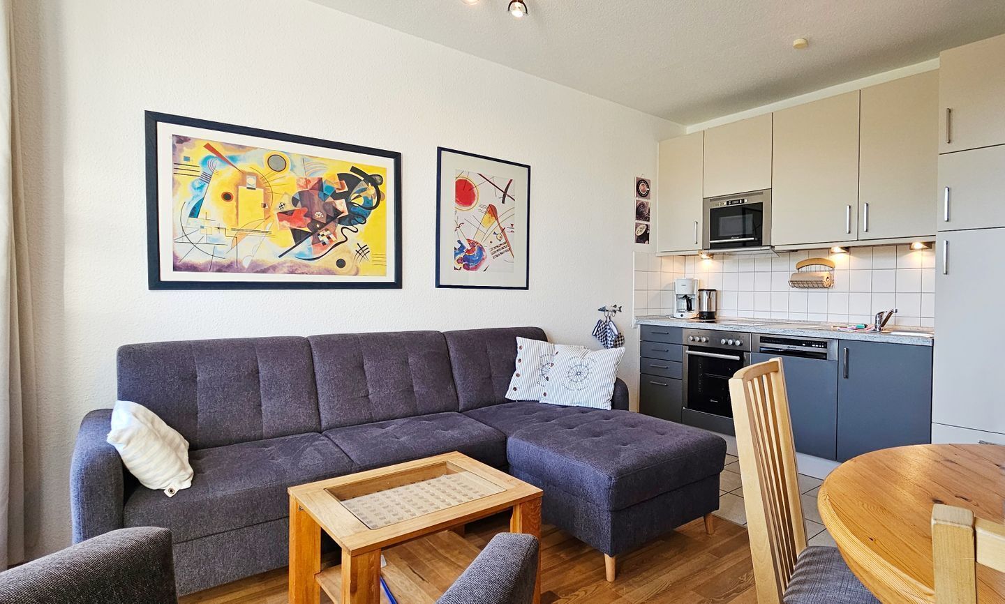Ein modernes Wohnzimmer mit einer grauen Couch, einem Holztisch und einem Essbereich mit Holztisch und Stühlen. Die Küche verfügt über weiße Schränke, eine Mikrowelle und einen Geschirrspüler. An der Wand hängen zwei abstrakte Gemälde.