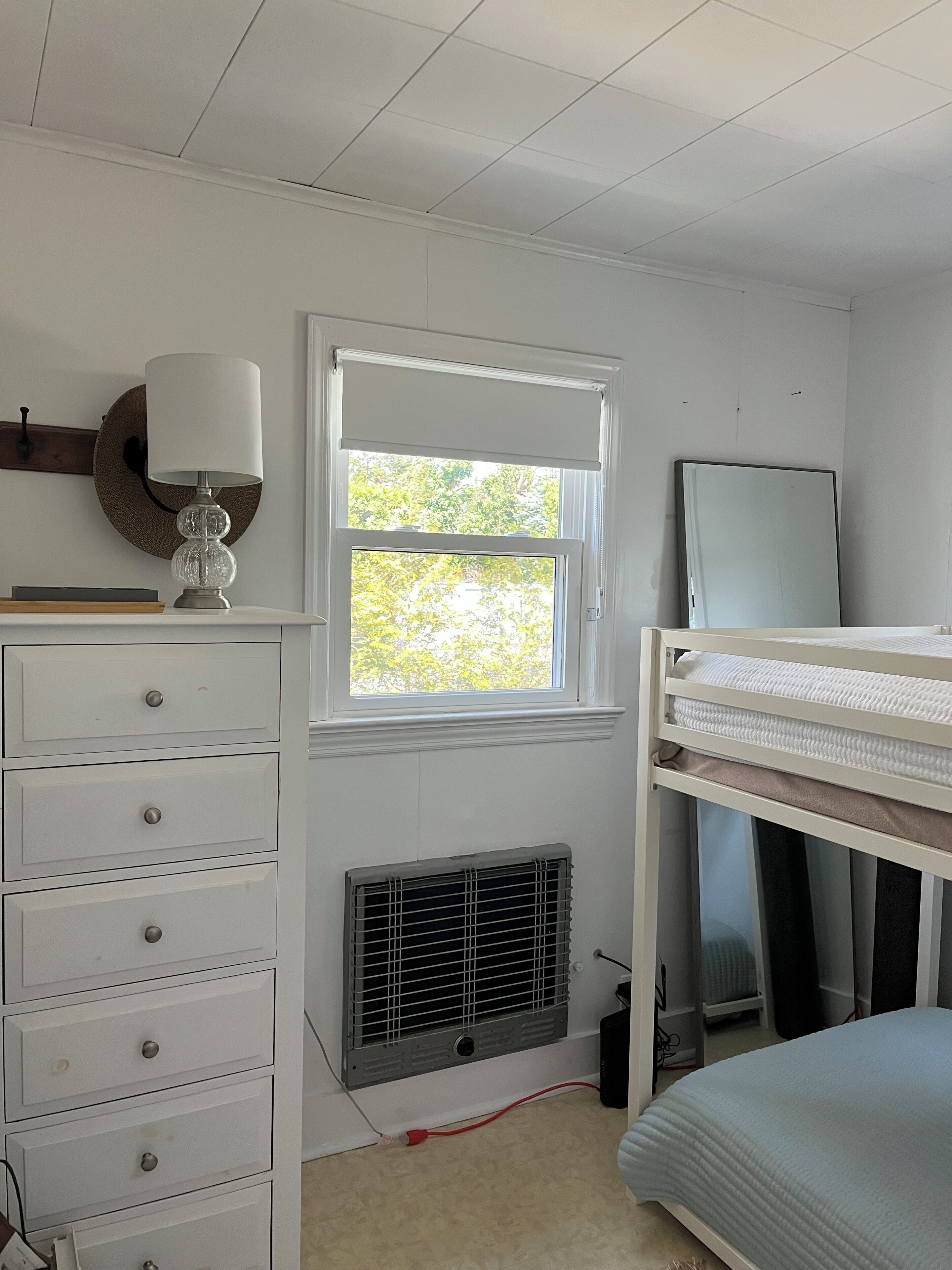 A bedroom with a bunk bed, a dresser, a lamp, a window with blinds, and a radiator.