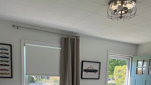 Un salon avec un canapé beige, deux poufs en osier, un tableau représentant une voiture et une porte donnant sur l’extérieur.