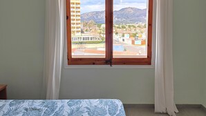 Una habitación con cama, ventana con cortinas y vistas a edificios y montañas.