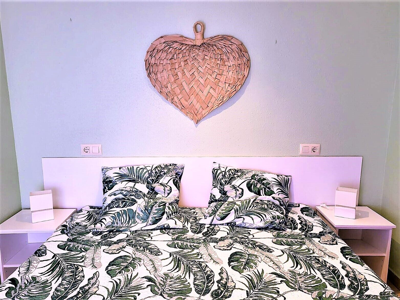 A bedroom with a bed, two nightstands, a heart-shaped wall decoration, and a floral-patterned bedspread.
