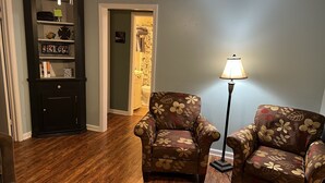 Una sala de estar con suelo de madera, dos sillones con estampado floral, una lámpara de pie, una estantería y una puerta que da a un baño.