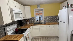 Una cocina con armarios blancos, microondas, estufa y refrigerador. Hay una ventana con vistas al exterior, y un reloj en la pared.