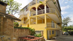 Un immeuble jaune à plusieurs étages, doté de balcons, d’un mur en pierre et d’un espace extérieur aménagé pour s’asseoir.
