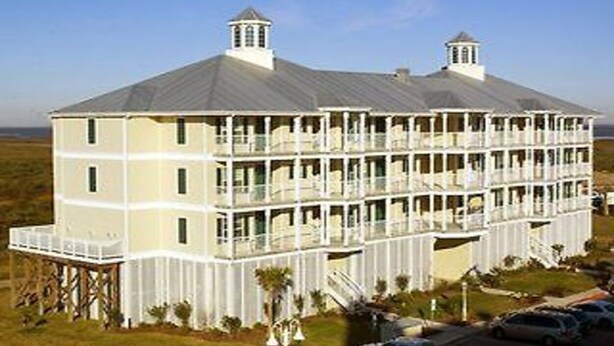 A large, multi-story building with balconies and a flat roof, situated in a grassy area with a clear sky.
