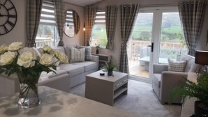 En rummelig stue med en grå sofa, et sofabord og et stort vindue med gardiner. Der er et spejl på væggen og et ur over sofaen.