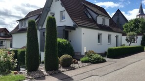 Ein zweigeschossiges Haus mit steilem Dach, einer kleinen angebauten Garage und einem gepflegten Garten mit Hecken und einem Weg.