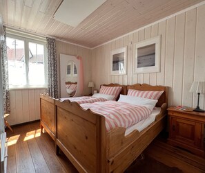 Ein Bett mit Holzboden, mit gestreifter Bettwäsche, ein Nachttisch mit Lampe und ein Fenster mit Vorhängen.