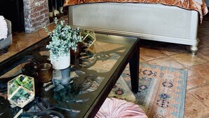 Un dormitorio con chimenea de ladrillo, una cama grande con muchos cojines, una mesa de centro de cristal con objetos decorativos y una alfombra estampada.