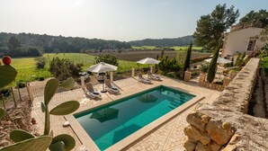 Una piscina rettangolare con bordo piastrellato, circondata da lettini e ombrelloni, con sullo sfondo campi verdi e alberi.