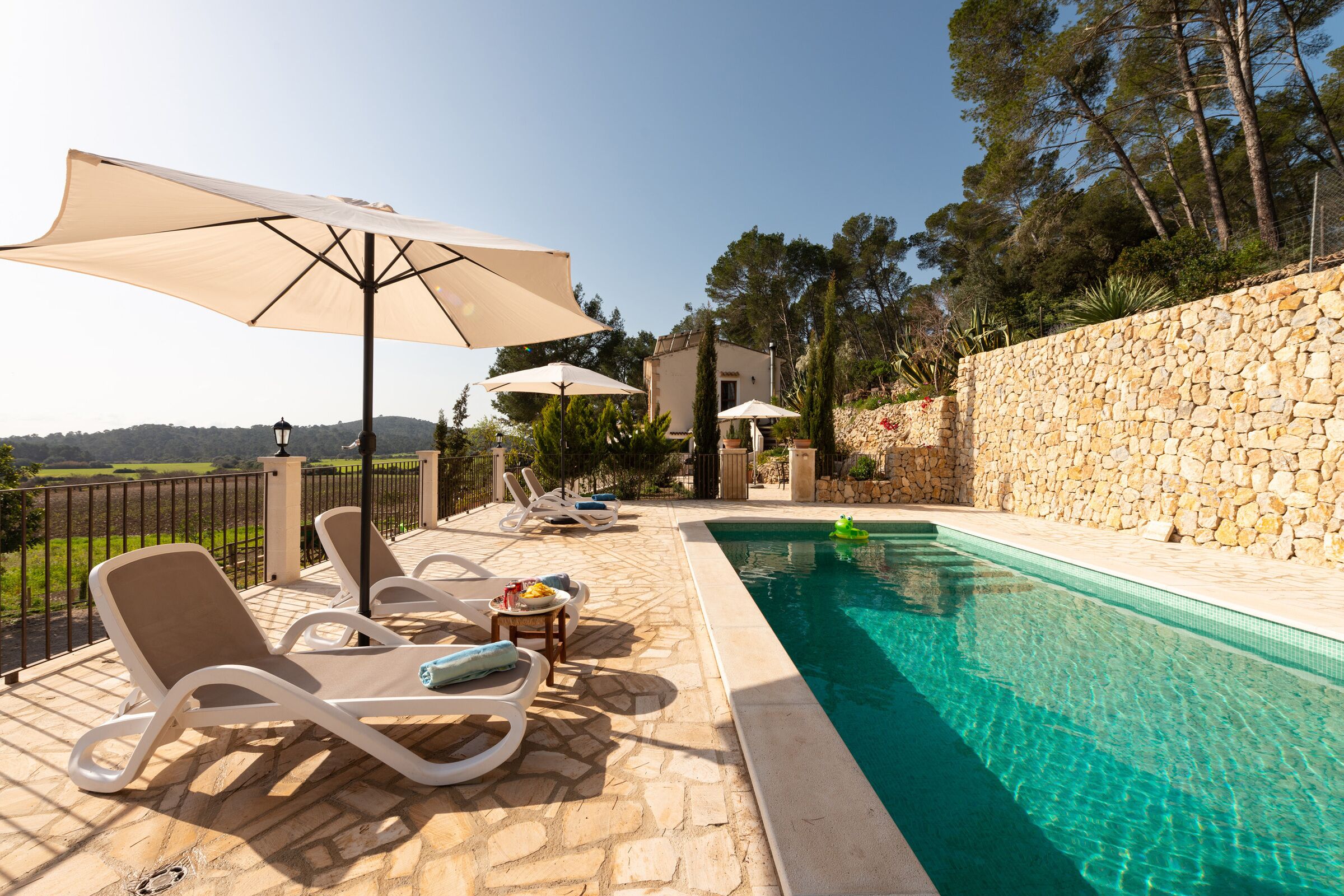 Un área junto a la piscina con sillones de descanso y sombrillas, un muro de piedra y vistas al campo.