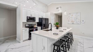 A modern kitchen with white cabinets, a large island with bar stools, stainless steel appliances, and a decorative wall piece. - The Modern/Luxe Vacation • Heated Pool •  Disney (Davenport)