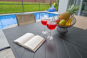 Mesa à beira da piscina com um livro aberto, duas bebidas vermelhas e uma cesta de frutas.
