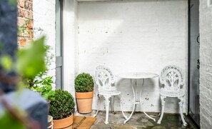 A small outdoor seating area with two white metal chairs and a table, surrounded by white walls and a brick wall. There are potted plants and a door in the background. - Bootham in Style -  a couples retreat that sleeps 3 guests  in 1 bedroom (York)