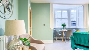 Ein Wohnzimmer mit beiger Couch, grüner Couch, einer Lampe und einer Pflanze. Im Hintergrund befindet sich ein Essbereich mit Tisch und Stühlen.