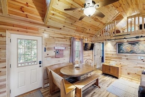 A wooden cabin interior with a dining table, chairs, a ceiling fan, a television, and a flag on the wall.
