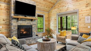 A cozy cabin interior with a stone fireplace, a flat-screen TV mounted above it, and a comfortable seating area with a sofa and armchairs. - Beyond Pines. Family-Friendly Escape & Retreat (Broken Bow)
