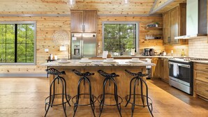 A spacious kitchen with wooden cabinetry, a central island with bar stools, stainless steel appliances, and large windows offering views of greenery. - Beyond Pines. Family-Friendly Escape & Retreat (Broken Bow)
