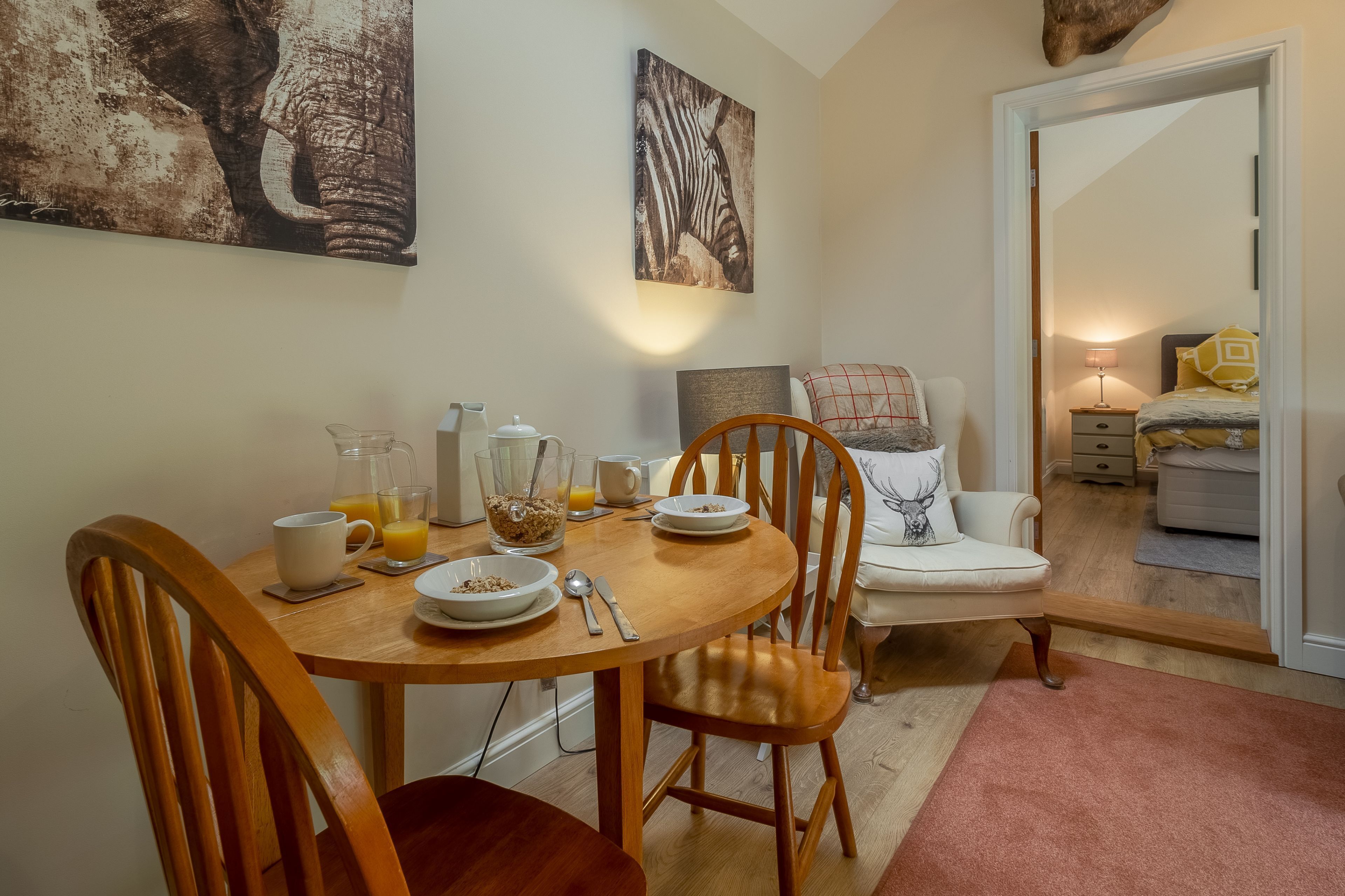 Un comedor con una mesa de madera dispuesta para el desayuno, un sofá con un cojín estampado y una cama al fondo.