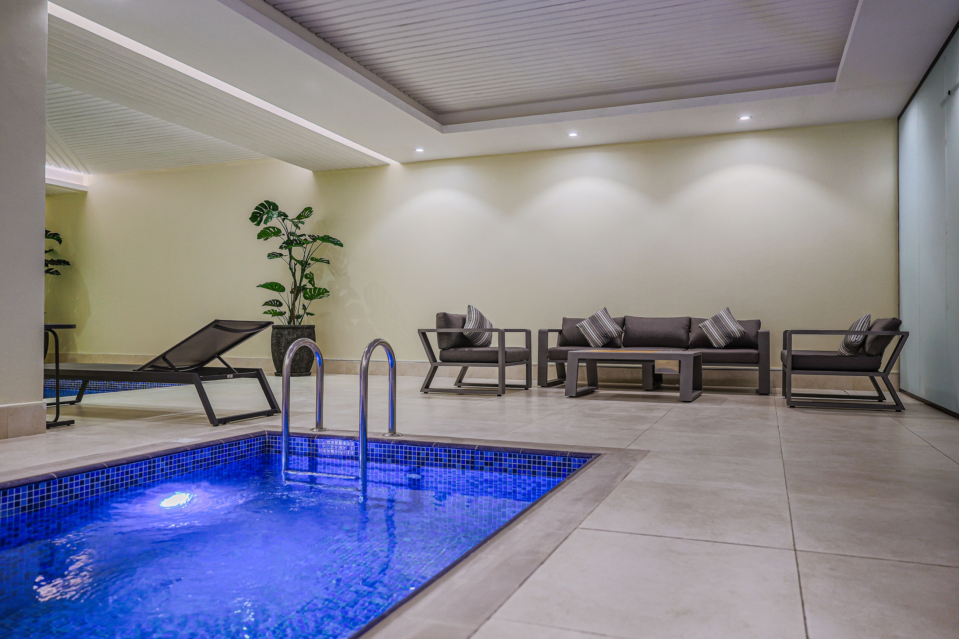 A modern indoor pool area with a tiled floor, lounge chairs, and a seating area with cushions.
