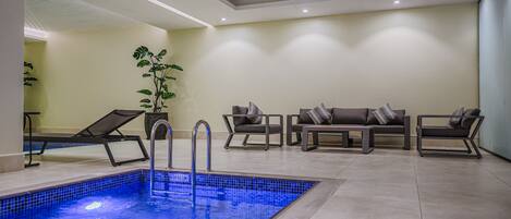 A modern indoor pool area with a tiled floor, lounge chairs, and a seating area with cushions.