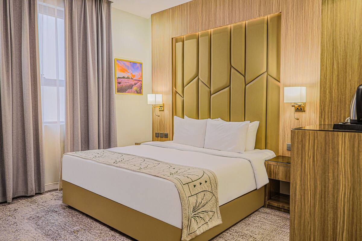 Una habitación de hotel con una cama grande, cabecero de madera, dos lámparas de noche, un escritorio y un cuadro en la pared.