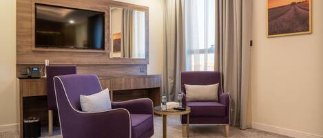A hotel room with two purple armchairs, a small round table, a TV mounted on a wooden panel, a desk with a computer, and a painting on the wall.