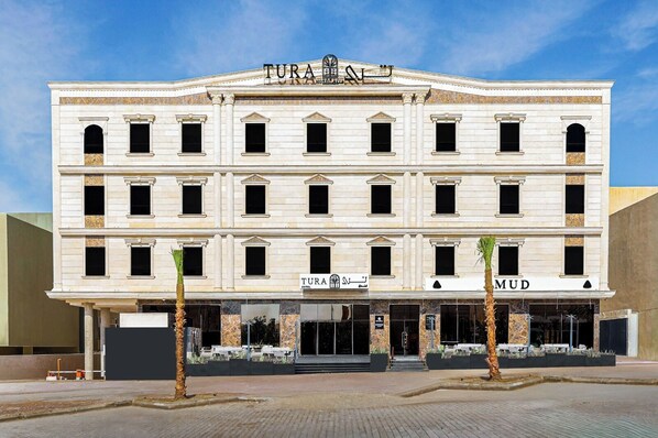 Un edificio de varios pisos con el nombre "TURA HOTEL" bien visible. Presenta un estilo arquitectónico clásico con columnas y una fachada simétrica.