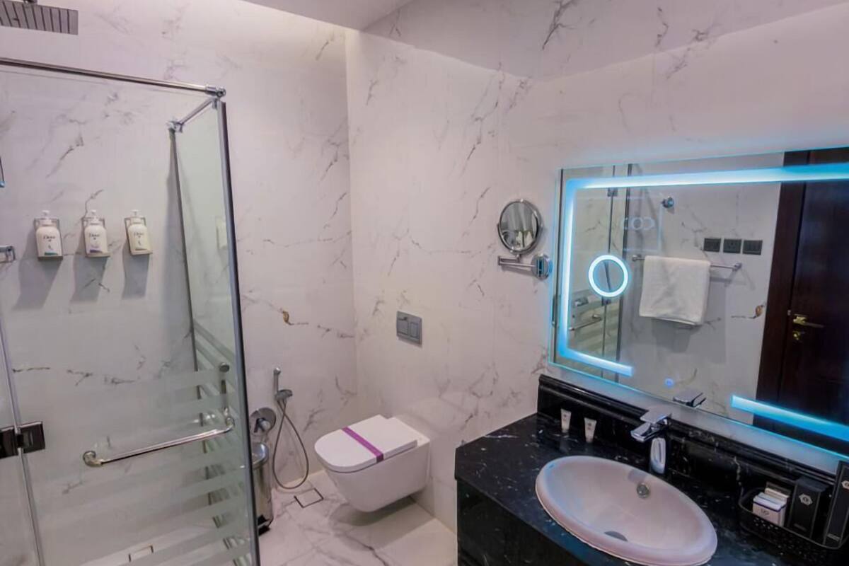 Un baño moderno con cabina de ducha de vidrio, inodoro blanco, lavabo negro con encimera de mármol y espejo con luz circular.