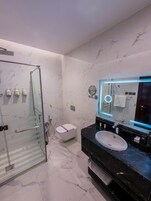Un baño moderno con cabina de ducha de vidrio, inodoro blanco, lavabo negro con encimera de mármol y espejo con luz circular.