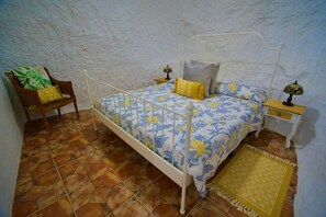 A bedroom with a white metal bed frame, floral bedding, a wooden chair with cushions, two bedside tables with lamps, and a patterned rug on a tiled floor.