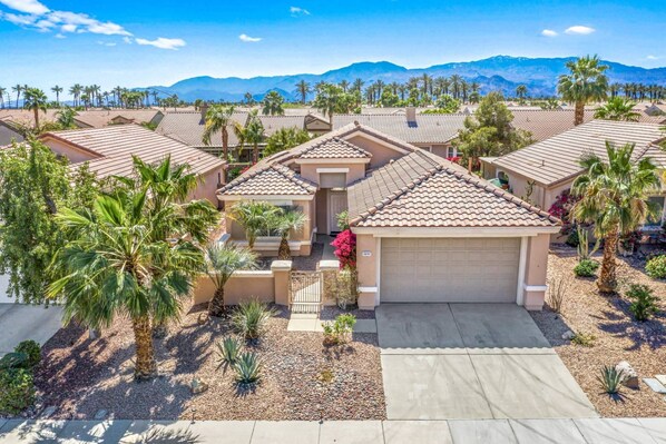 A residential area with multiple houses featuring similar architecture, surrounded by palm trees and a mountainous backdrop.