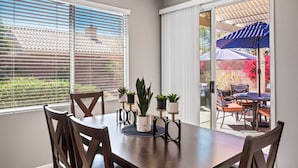 Un comedor con una mesa dispuesta para cuatro personas, rodeada de sillas de madera. Hay plantas en macetas sobre la mesa y una puerta corrediza de vidrio da a un patio exterior con una sombrilla azul.