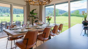 A dining area with a long table set for six, surrounded by chairs, with a chandelier overhead and a view of a green landscape through large windows. - Detached luxury chalet (Kreischberg)