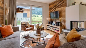 A modern living room with a sofa, armchair, and coffee table, a flat-screen TV mounted on a wooden wall, and a bookshelf with books and decorative items. - Detached luxury chalet (Kreischberg)