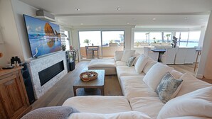 Un salón amplio con un sofá blanco, una chimenea, un televisor grande y vistas al océano a través de amplios ventanales.