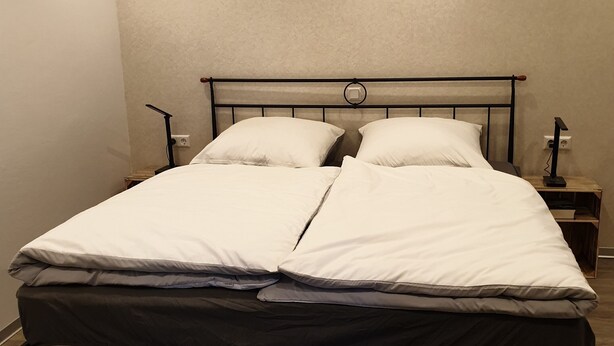 Una cama doble con estructura metálica y ropa de cama blanca, una mesita de noche con lámpara, y una lámpara de pared.