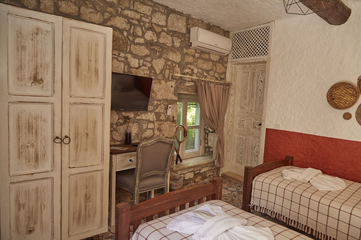 A room with stone walls, a wooden desk, a chair, a bed with a checkered blanket, and a television.