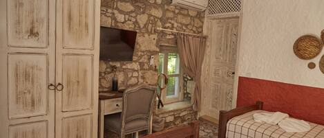 Una habitación con paredes de piedra, un escritorio de madera, una silla, una cama con manta a cuadros y un televisor.