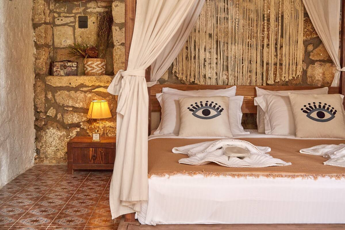 A bedroom with a stone wall, a wooden nightstand with a lamp, a bed with a canopy, and decorative pillows.
