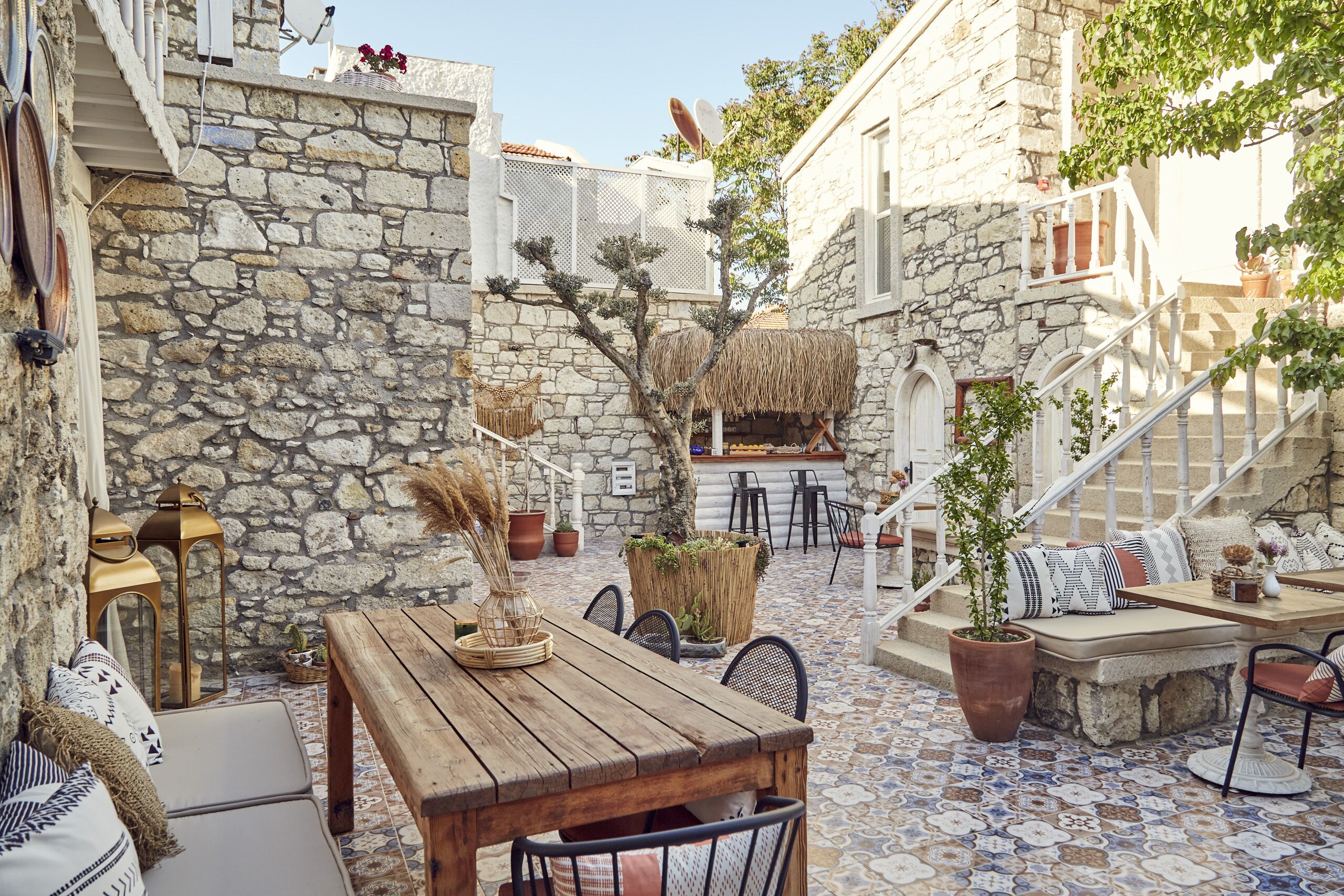 A stone-walled courtyard with a wooden table and chairs, potted plants, and a staircase leading to an upper level.