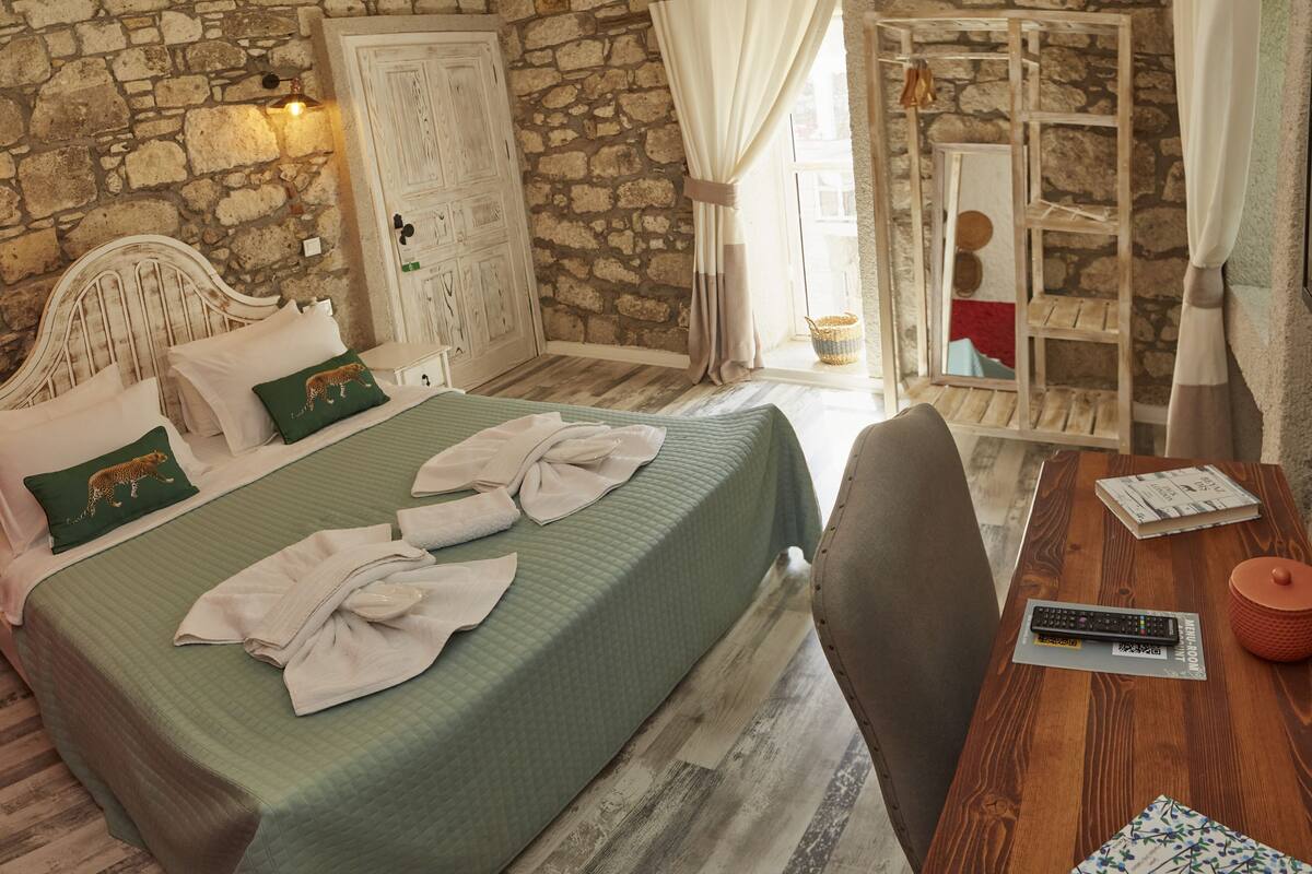 A room with a bed, a wooden table, a mirror, and a stone wall.